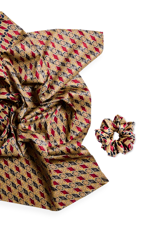 A luxury silk accessory set featuring a large scarf and a matching mid-size scrunchie with an intricate geometric diamond pattern in burgundy, gold, and black tones on a white background.