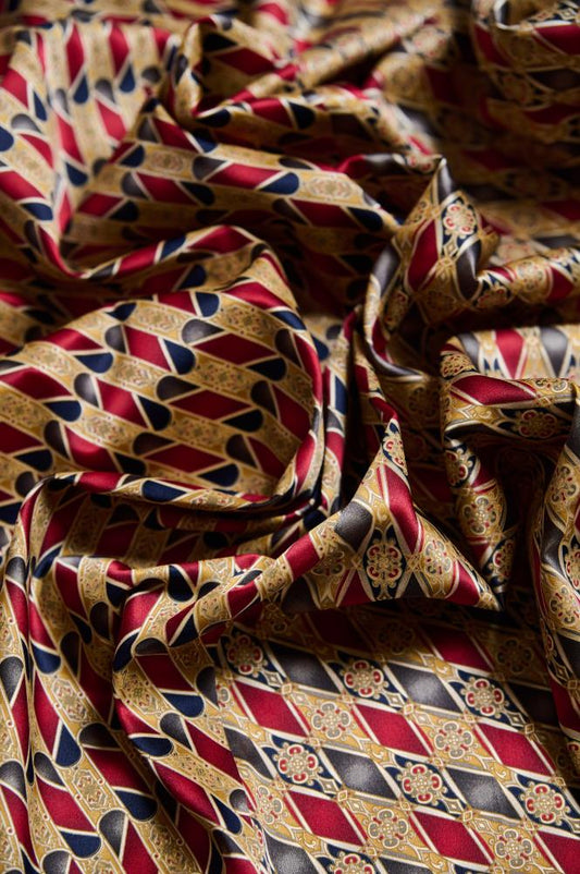 A close-up of crumpled silk fabric with a bold geometric pattern. The fabric intertwines rich red, gold, and black colors, forming sequences of diamonds and stylized ornaments. The glossy silk surface and deep folds create a sense of luxury and dynamism, highlighting a classic design with a touch of oriental motifs.