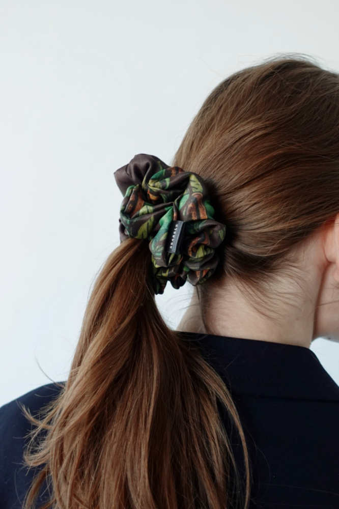 A back view of a girl with brown hair tied in a ponytail. Her hair is styled with a large, voluminous dark brown fabric scrunchie featuring green and orange botanical patterns. She is wearing a dark blazer, and the background is light and neutral.