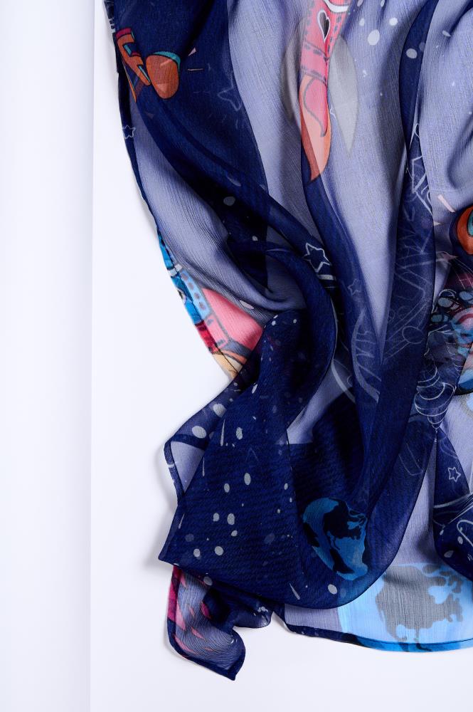 A close-up photo showing the folds of a dark blue, sheer, and lightweight scarf against a white background. The fabric is decorated with colorful space-themed illustrations, including rockets, stars, and planets. The photo highlights the delicate transparency of the fabric, the soft drapes, and the vibrant colors of the pattern, creating a sense of lightness and creativity.