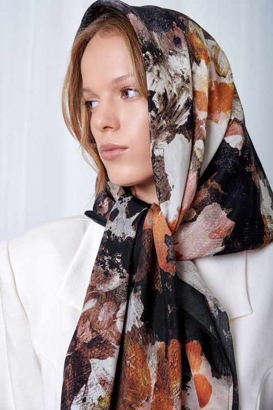 Woman wearing a floral patterned headscarf and white blazer, gazing to the side. The scarf features earthy tones, creating an elegant and thoughtful look.