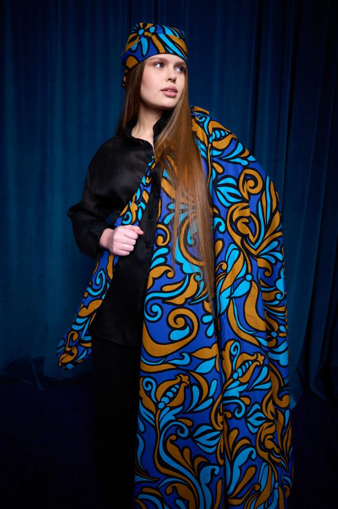A young woman standing against dark blue curtains, wearing black clothing. She is wrapped in a large, vibrant blanket and wearing a matching headscarf. Both accessories feature a rich Baroque-style pattern in shades of royal blue, cyan, and bronze. Her long brown hair falls over her shoulders, accentuating the texture of the accessories.