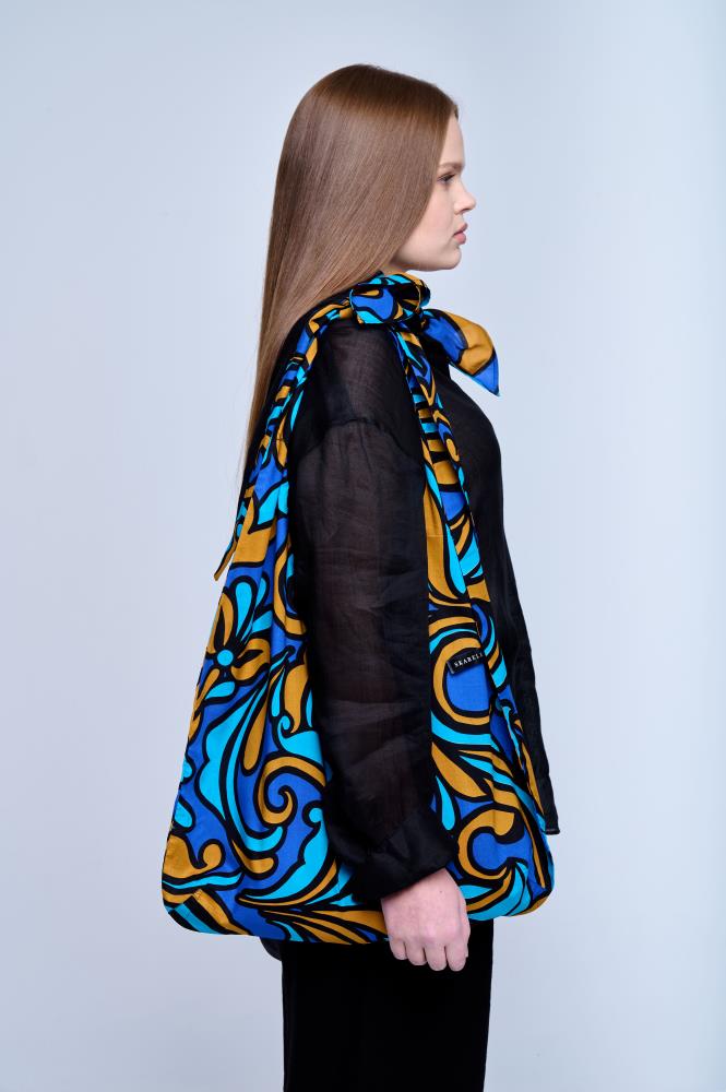A young woman standing in profile against a light background, wearing a black shirt. She is carrying a large fabric tote bag on her shoulder, featuring a vibrant Baroque pattern in shades of royal blue, cyan, and bronze. The bag's straps are tied in a knot at the top, giving the accessory a unique shape.