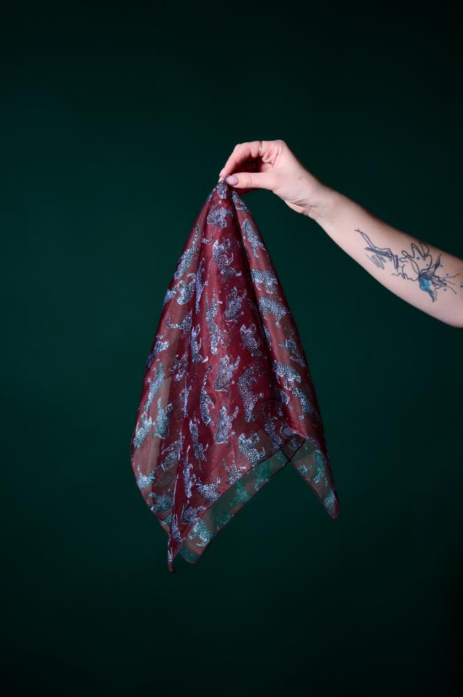 A hand holding a burgundy silk scarf with a stylish leopard pattern against a dark green background. The photo shows a tattooed arm holding the light, sheer fabric, which features a subtle sheen and grey-toned animal motifs.