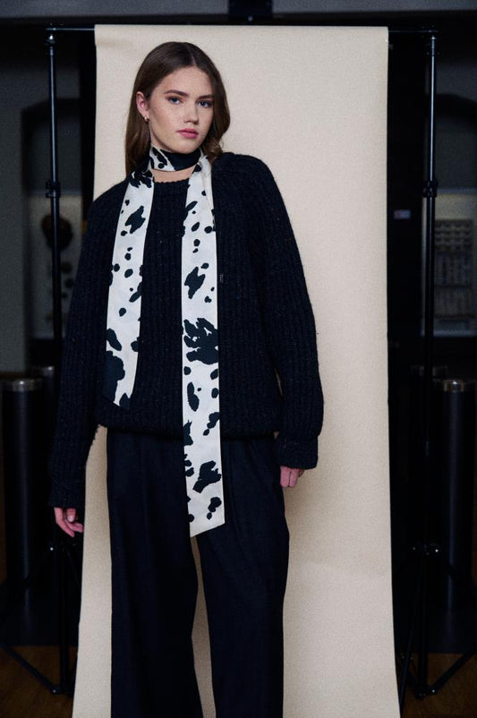 A young woman poses in a studio in front of a light backdrop. She is wearing a chunky knit dark sweater and wide black trousers. A long, narrow silk scarf is stylishly wrapped around her neck, featuring a bold black and white pattern reminiscent of Dalmatian spots or abstract blotches. The ends of the scarf hang freely in front, adding a contrasting and modern accent to the dark outfit.