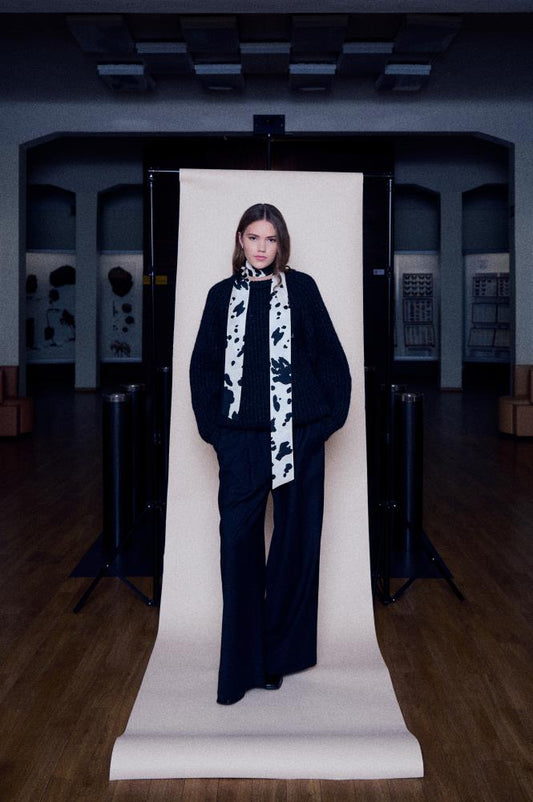 A full-length photo of a young woman posing against a studio backdrop set up in a spacious, dark hall with wooden floors. She is wearing a black chunky knit sweater and wide black trousers, with a long, narrow silk scarf featuring contrasting black spots on a white background wrapped around her neck. The light backdrop roll creates a sharp vertical accent in the dark interior, highlighting the model's silhouette and stylish, minimalist outfit.
