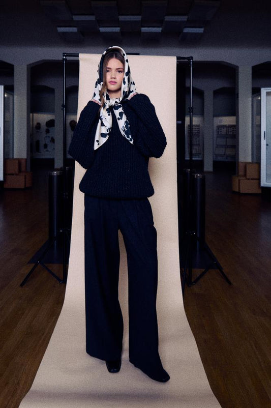 A full-length photo of a young woman posing against a studio backdrop set in a large, dark hall with wooden floors. She is wearing a black chunky knit sweater and wide black trousers, with a silk scarf wrapped around her head. The scarf features a bold black and white pattern with irregular spots and is stylishly tied under her chin.