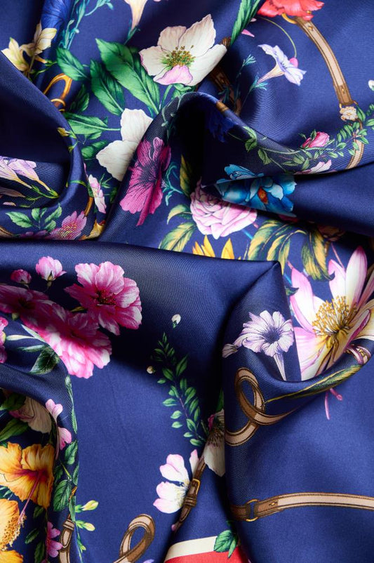 A close-up of crumpled, glossy silk fabric with a rich floral pattern scattered across a dark blue background. The fabric features pink, white, yellow, and purple blossoms along with green leaves, intertwined with brown leather strap motifs. The soft play of light on the folds of the fabric emphasizes the material's luxury, smoothness, and the detail of the pattern.