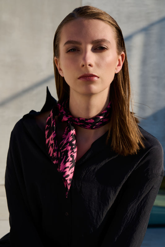 A model poses in a fashionable look with a silk scarf stylishly tied around her neck. The scarf features a vibrant black and pink graphic pattern that stands out in contrast against a dark shirt. Natural lighting and a minimalist setting emphasize the modern aesthetic and elegant style of the accessory.
