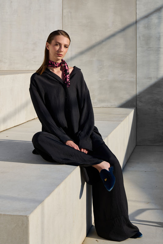 A model sits on concrete stairs in an industrial setting, wearing a loose black suit and a vibrant silk "Twilly" scarf. The scarf is black and pink with a graphic pattern, stylishly tied around her neck, adding a colorful accent to the minimalist look. Blue velvet shoes and sun shadows complement the modern and elegant composition of the photo.