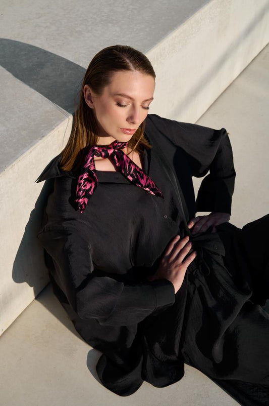 A model poses leaning against a concrete wall, wearing a loose black suit and a vibrant silk scarf. The scarf is black and pink with a pattern, stylishly tied around her neck as a central accent. Natural sunlight highlights the fabric textures and the modern, minimalist aesthetic of the photo.