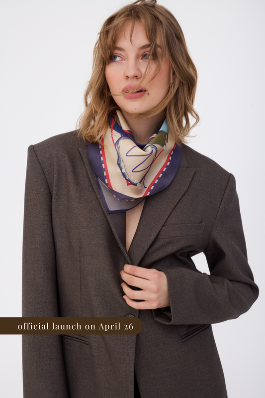 A woman wearing an oversized brown blazer and a silk neck scarf with an abstract colorful pattern. The scarf features a navy blue border with red and white geometric details. A gold banner at the bottom reads "official launch on April 26". The photo is set against a clean white background.
