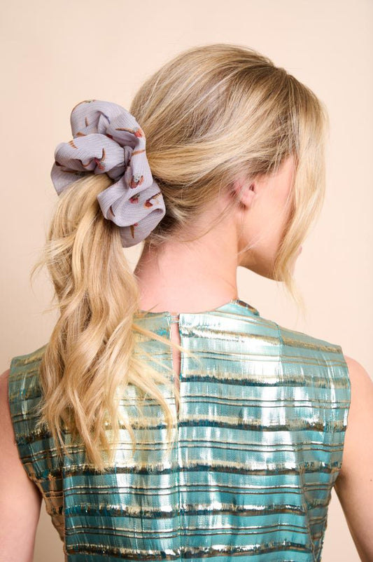 A back view of a woman with blonde, wavy hair tied in a medium-height ponytail. Her hair is styled with a large, voluminous greyish silk scrunchie featuring small ice cream patterns. She is wearing a shiny blue and gold striped top, set against a light, neutral background.