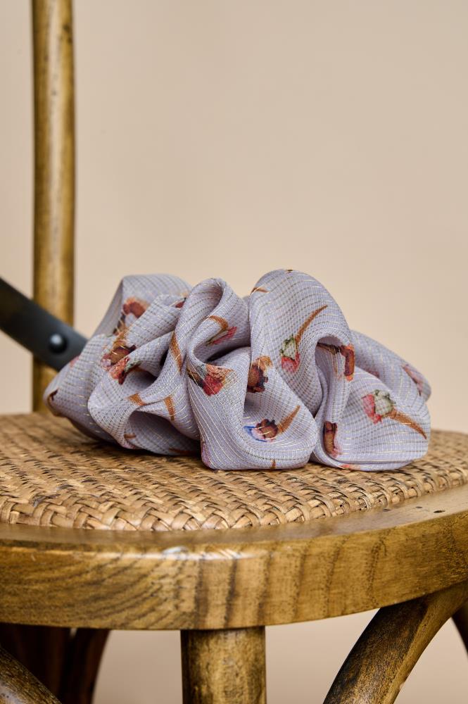 A voluminous grey silk scrunchie placed on the woven seat of a wooden chair. The accessory features small ice cream patterns and a subtle metallic sheen in the fabric. The warm lighting and natural wood textures create a cozy, rustic atmosphere.