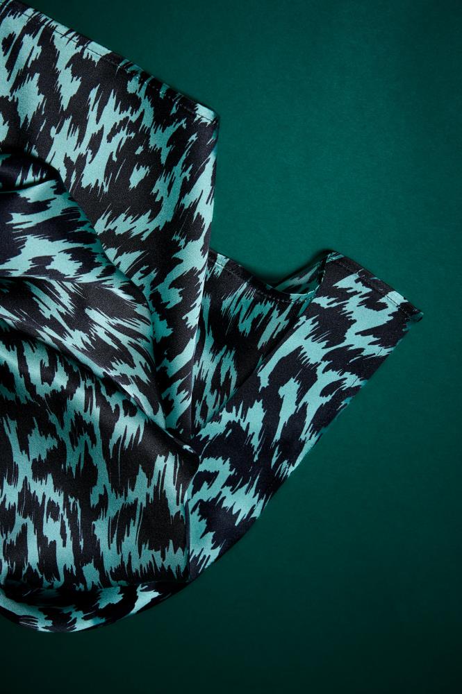 A detail of a teal and black patterned silk scarf against a dark green background. The photo highlights the softness of the fabric, the subtle silk sheen, and the dynamic graphic pattern. This composition emphasizes the accessory's luxury, quality texture, and elegant modern design
