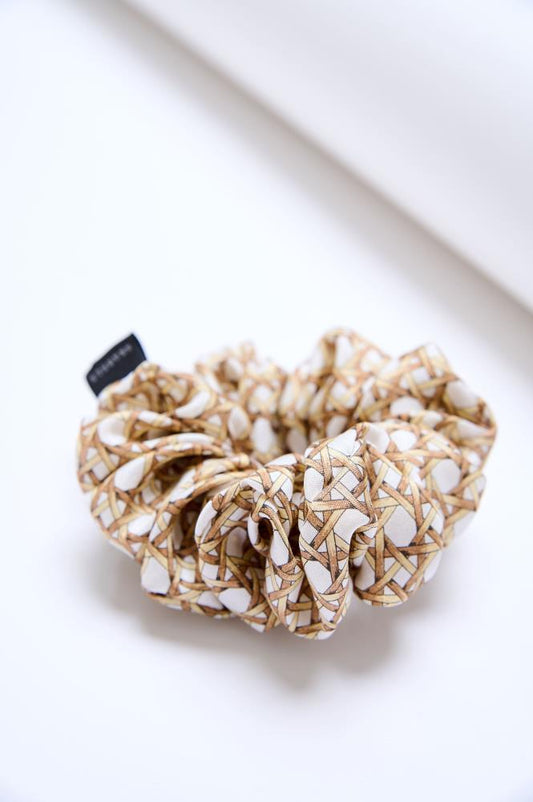 A large, voluminous fabric scrunchie placed on a light, neutral surface. The accessory features a cream-colored fabric with brown woven patterns, resembling wicker or basket weaving. A small black brand label is visible on the left side.