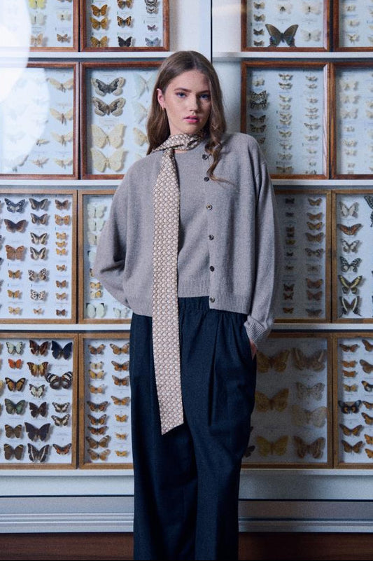 A model posing with a skinny silk scarf with a beige geometric pattern, paired with a taupe knit cardigan and dark high-waisted trousers, standing in front of framed butterfly collections.