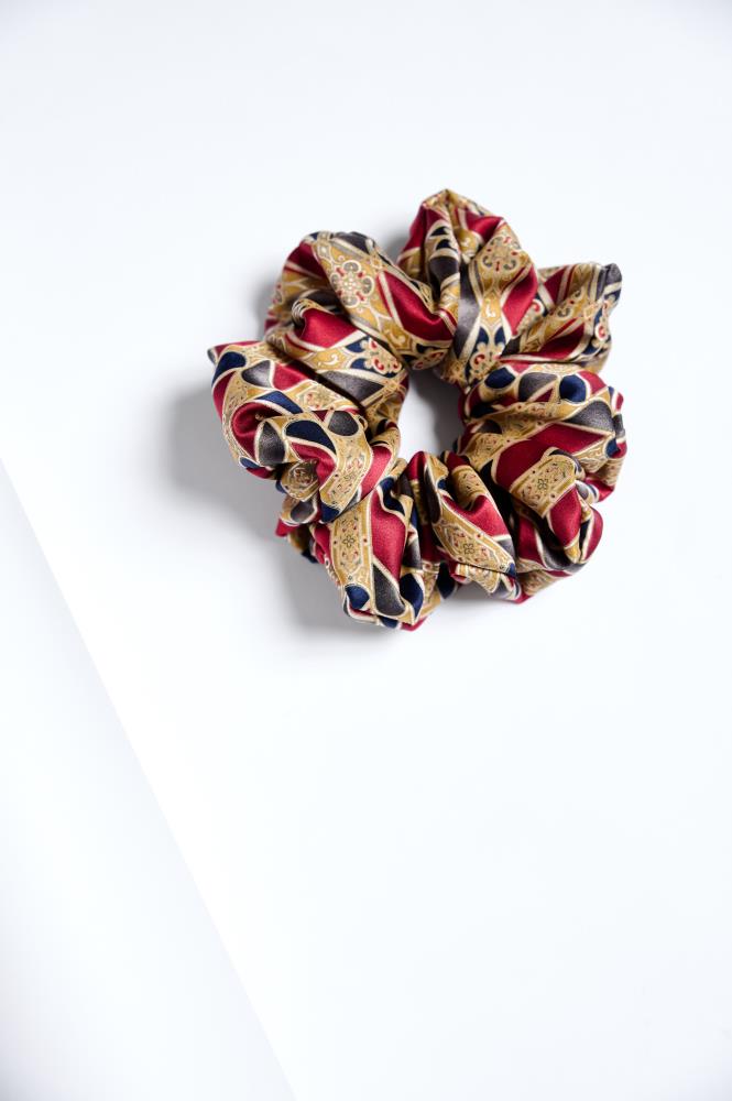 A large, voluminous fabric scrunchie placed on a clean white background. The accessory features a luxurious, detailed pattern with burgundy, gold, and navy blue motifs, reminiscent of classic ornamental style. The fabric's sheen and folds create a sense of depth and elegance.