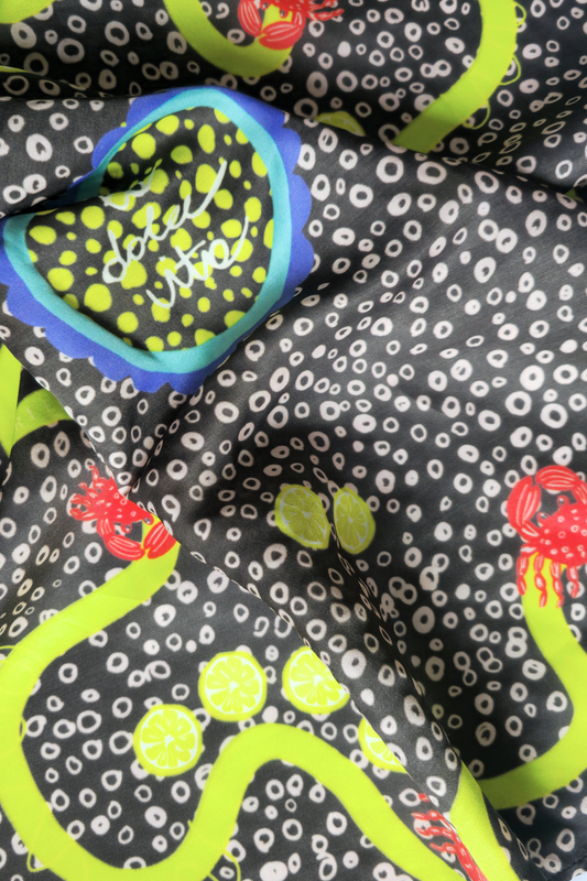 Macro close-up of draped Skarele La Dolce Vita viscose & silk blend scarf. Shows premium satin texture, white dotted print on dark greyish-brown fabric, lime green wavy lines, crabs, and lemon illustrations.