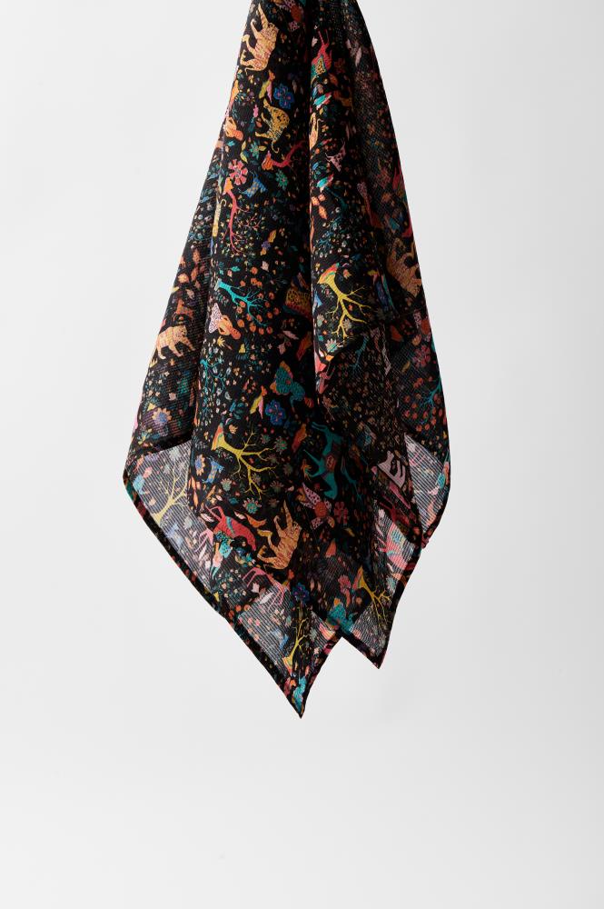 A colorful scarf with a vibrant abstract pattern featuring plant and animal motifs in yellow, red, and teal on a black background, hanging elegantly.