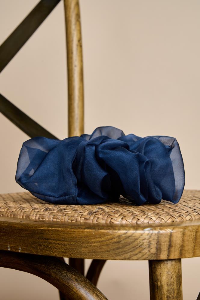 A voluminous, dark blue sheer fabric scrunchie placed on the woven seat of a wooden chair. The light and abundant fabric folds give the accessory a luxurious, airy look. The background is warm and neutral, and the texture of the wooden chair creates a cozy atmosphere.