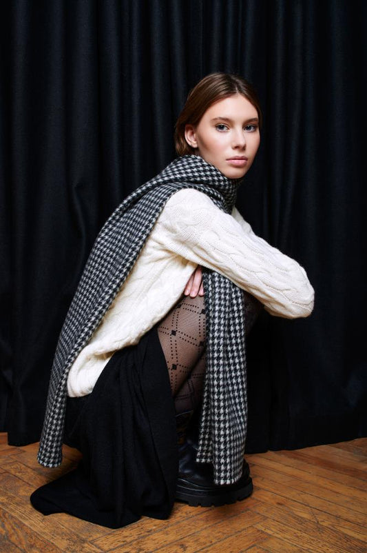A stylish woman poses in a crouching position against a backdrop of black curtains on a wooden floor. She is wearing a light cable-knit sweater, a dark skirt, patterned tights, and chunky black boots. A long wool scarf with a classic black and white houndstooth pattern is draped over her shoulder and wrapped around her neck. Her calm gaze is directed toward the camera, and the composition highlights a cozy, autumnal outfit.