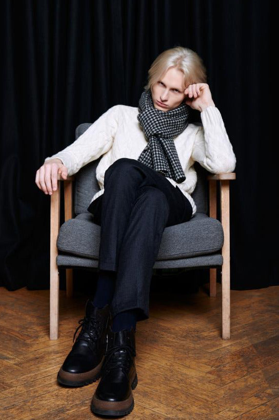 A young blonde man sits in a grey armchair against a backdrop of dark curtains. He is wearing a light cable-knit sweater, dark trousers, and chunky black boots. A wool scarf with a classic black and white houndstooth pattern is stylishly wrapped around his neck. The man is sitting with his legs crossed, leaning his head on one hand, looking toward the camera. The composition creates a calm, modern, and cozy atmosphere.