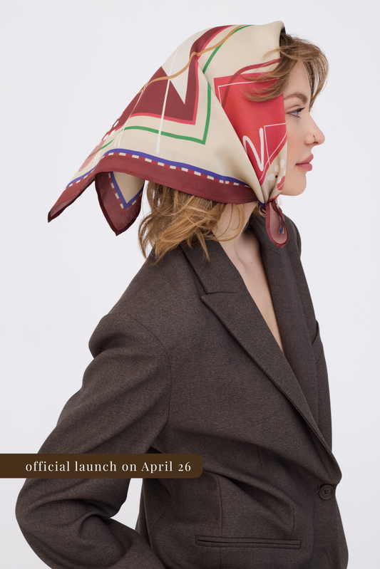 Model showcasing a patterned silk scarf worn over the head and knotted under the chin. The accessory has a cream base with large abstract graphic shapes in warm tones and a Chestnut Brown border. The shot highlights the contrast between the smooth silk and the textured brown jacket.