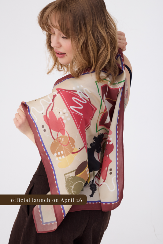 A woman viewed from the back, holding up a large silk scarf to showcase its full pattern. The scarf features an abstract artistic design with terracotta, ochre, and olive green shapes on a cream background, finished with a deep burgundy border. She is wearing dark brown trousers against a white studio backdrop.