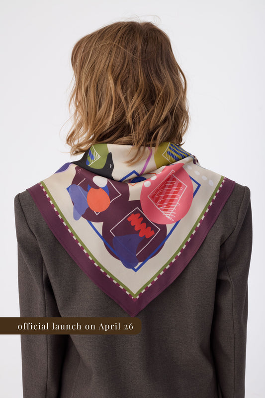 rear view of a woman with curly brown hair wearing a dark brown tweed blazer. A silk scarf with an abstract artistic pattern featuring blue, orange, and red shapes is tied around her neck, draped in a point. The scarf has a deep burgundy border. A gold banner at the bottom reads "official launch on April 26". The background is a clean white studio setting.