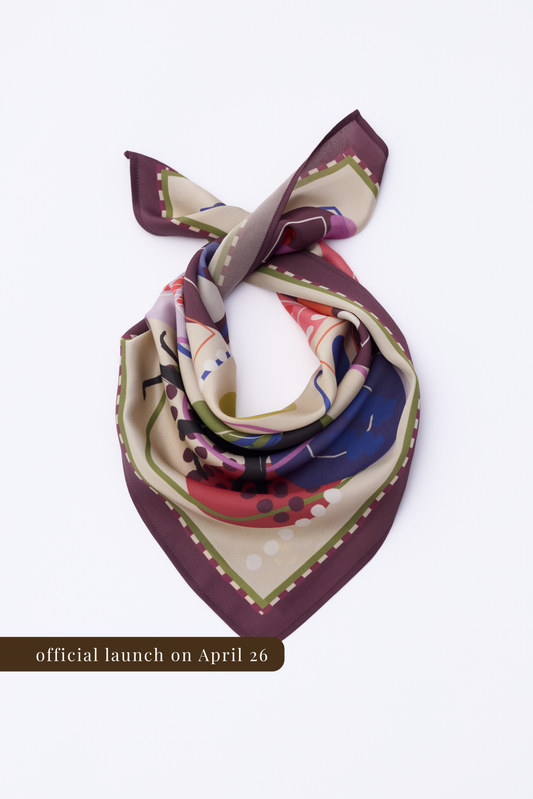 Abstract patterned silk scarf with a burgundy hem, laid out on a white surface. The image emphasizes the print's colors and the fluid drape of the fabric. Includes a text banner for the April 26 official launch.