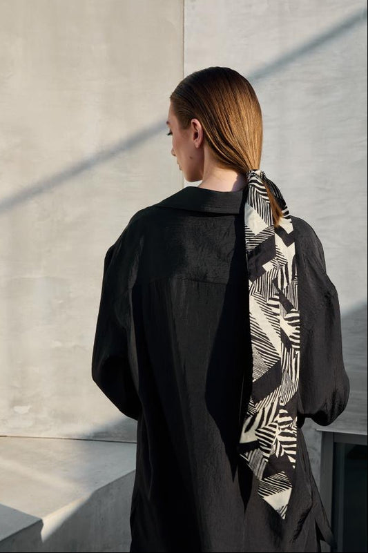 A back view photo showing a stylish woman's look with a narrow silk scarf tied into a low ponytail. The scarf features a graphic black and white botanical pattern that stands out contrastingly against a dark shirt and a concrete wall background. A modern hair accessory that adds elegance and bohemian charm to an everyday look.