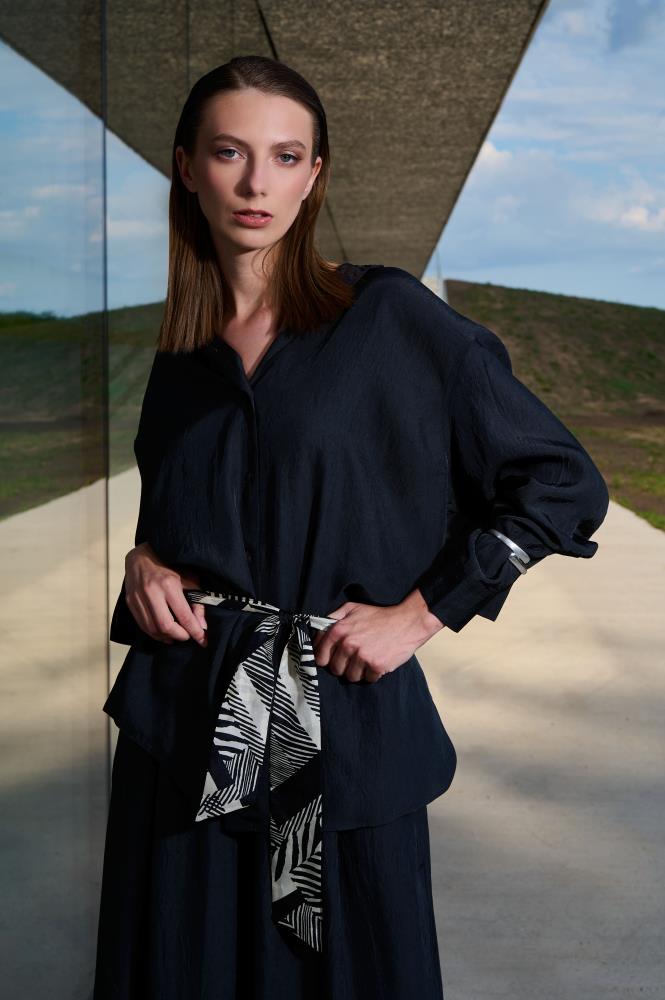 A model poses outdoors by a glass wall, wearing a dark shirt accessorized with a narrow "Twilly" silk scarf used as a stylish belt. The scarf features a graphic black and white pattern that highlights the waist and adds an elegant accent to the modern look. The industrial setting and natural lighting emphasize the minimalist aesthetic of the accessory.
