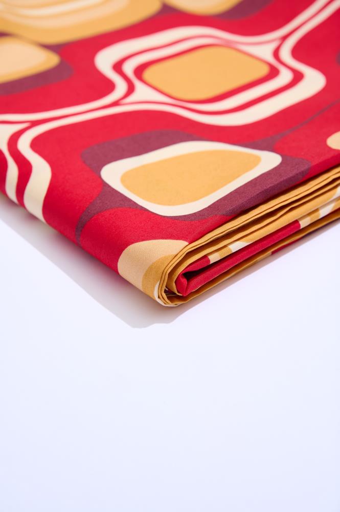 A close-up of folded fabric against a clean white background. The material features a bold retro-style pattern with rounded squares in yellow, orange, red, and burgundy. This detailed shot highlights the texture and the richness of the fabric's design.