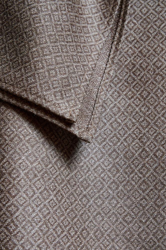 A close-up of a dark grey or brown fabric with a fine, repeating diamond-shaped geometric pattern. The photo shows the corner of a scarf or shawl and the stitching along the edge. The fabric's texture appears soft and dense, while the pattern adds subtle depth and elegance to the material.