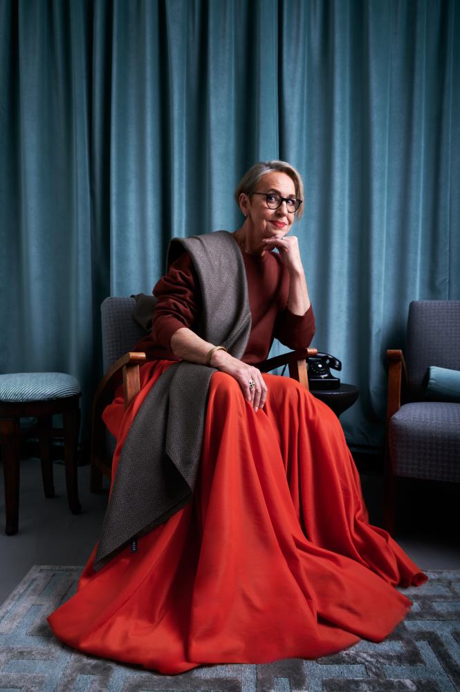 A portrait of an older woman with short hair and glasses sitting in an armchair against a backdrop of blue velvet curtains. She is wearing a burgundy sweatshirt and bright red maxi skirt that drape in rich folds onto a grey patterned rug. A stylish grey-brown wool scarf is draped over her shoulder, and she rests her chin on one hand while looking at the camera. The composition is completed by a retro black telephone on a side table, creating an elegant and peaceful atmosphere.