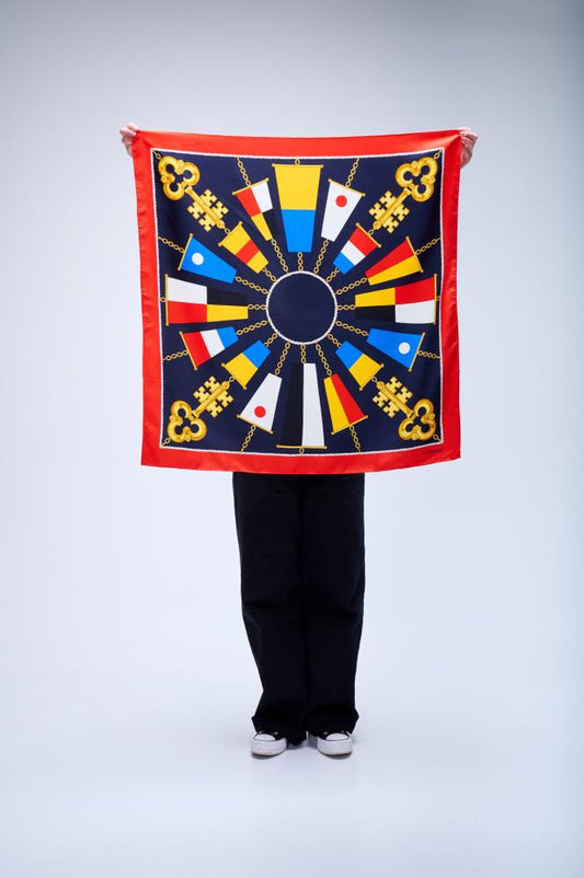 A person holding up a large square silk scarf with a nautical theme, featuring gold keys and maritime flags arranged in a sunburst pattern on a navy blue background with a red border.