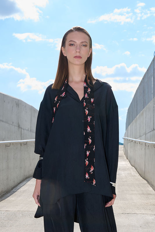 A model poses in a fashionable, industrial setting between concrete walls, looking into the camera. She is wearing dark moss grey fabric shirts, decorated with a stylish silk tie-scarf featuring a vibrant pink flamingo pattern on a black background. Her look emits a modern, elegant, and slightly playful mood.