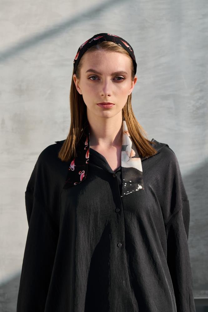 A model poses in a fashionable look with a narrow "Twilly" silk scarf. The scarf is black, decorated with a small, repeating pink flamingo pattern and blue accents, stylishly tied as a headband and drapes over a dark shirt. Soft lighting highlights the silk texture and pattern detail, emphasizing this playful and elegant accent.