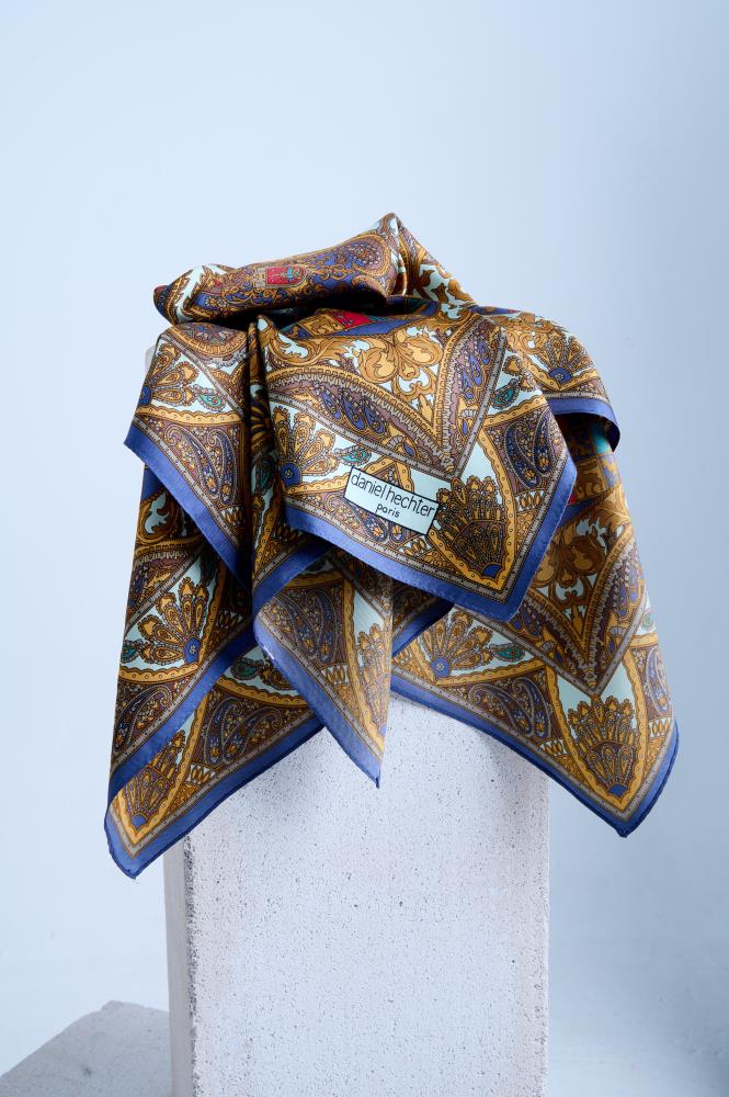 A silk scarf with an intricate heraldic and paisley print in gold, bronze, and light blue, artfully draped over a white stone pedestal in a professional studio setting.