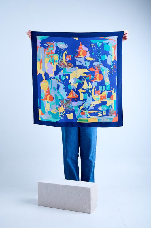 A person holding up a large square navy blue silk scarf featuring a vibrant, abstract underwater-themed print with colorful fish, coral-like shapes, and surreal elements in orange, yellow, and turquoise, framed by a dark blue border.