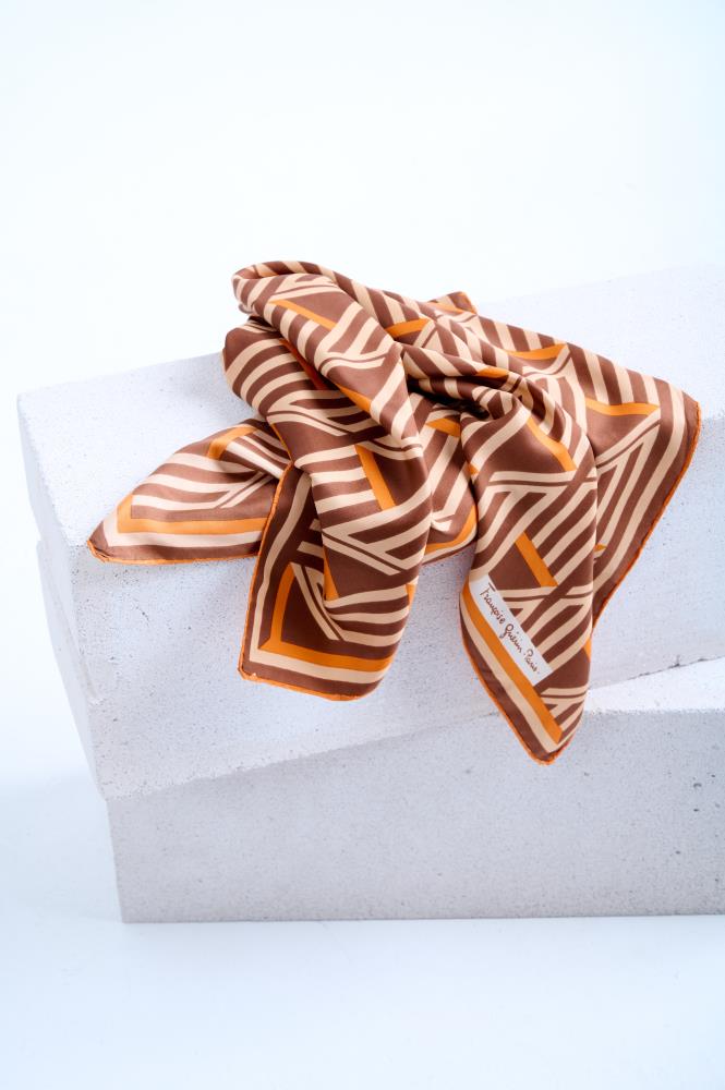 A vintage silk scarf featuring a bold brown, cream, and orange geometric striped pattern, artfully draped over a white textured stone pedestal in a bright studio setting.