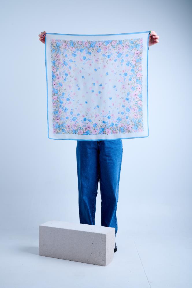 A person in blue jeans holding up a large square semi-transparent silk scarf featuring a delicate pastel floral print with blue and pink blossoms, framed by a thin light blue border, against a neutral studio background.