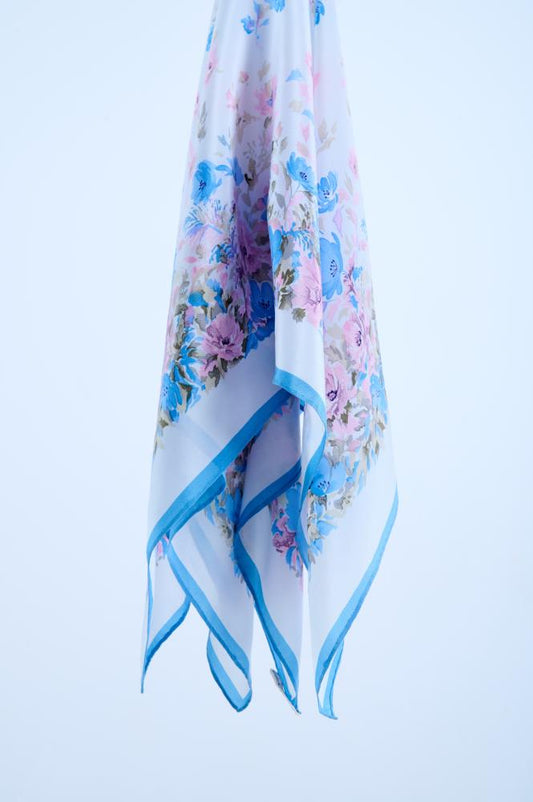 A lightweight, semi-transparent white silk scarf featuring a delicate floral print with bright blue and pink blossoms, elegantly draped to showcase its fluid texture and thin blue border against a clean studio background.