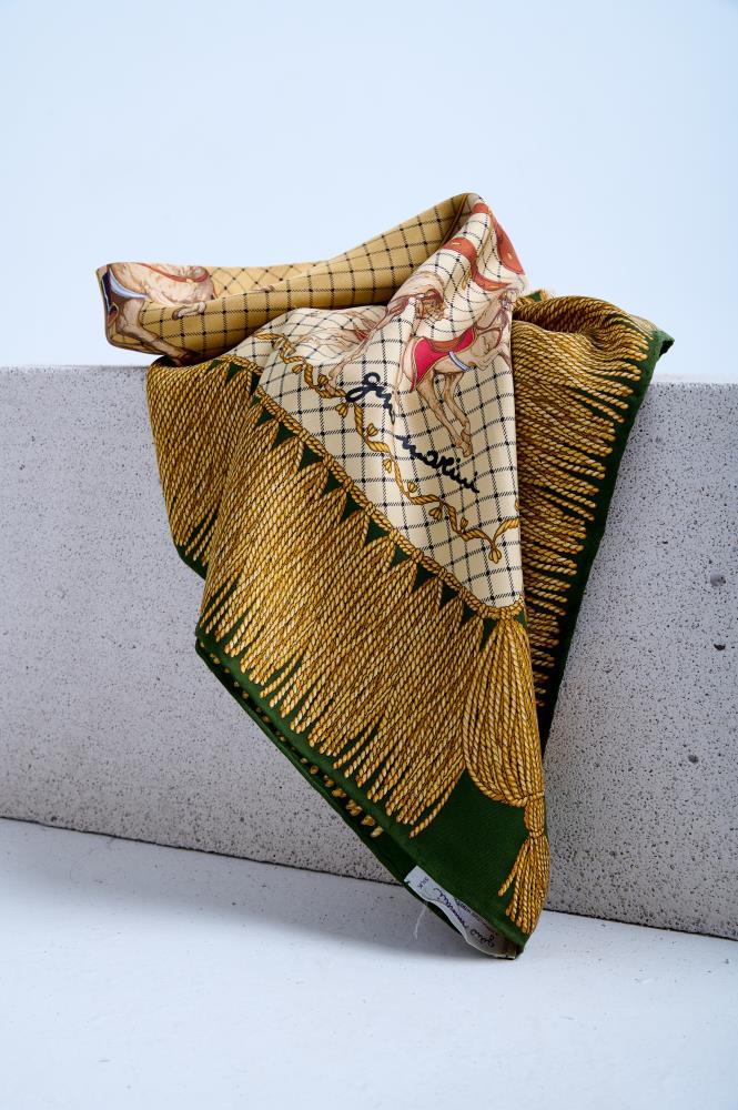 A luxury silk scarf with a vintage equestrian theme, featuring illustrated horses and a signature, artfully draped over a white stone block. The design includes a prominent gold tassel fringe pattern on a deep green border.