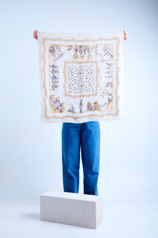 A person in blue jeans holding up a large square cream silk scarf, revealing a complete vintage-style design with ornate gold filigree borders and detailed violet illustrations of figures and landscapes against a neutral studio background.