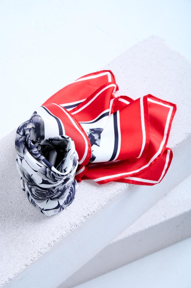A luxury silk scarf with a bold red, white, and navy blue striped border and a black and white floral rose print, artfully bunched and draped over a textured white stone pedestal in a studio setting.