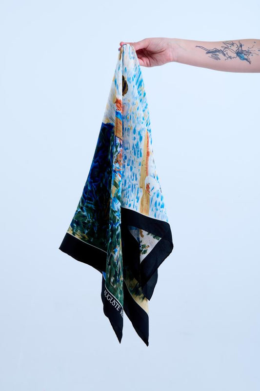 A tattooed hand holds up a luxury silk scarf featuring a vibrant art print inspired by Auguste Renoir's paintings, with impressionistic scenes of nature and figures in blue, green, and cream, framed by a bold black border.