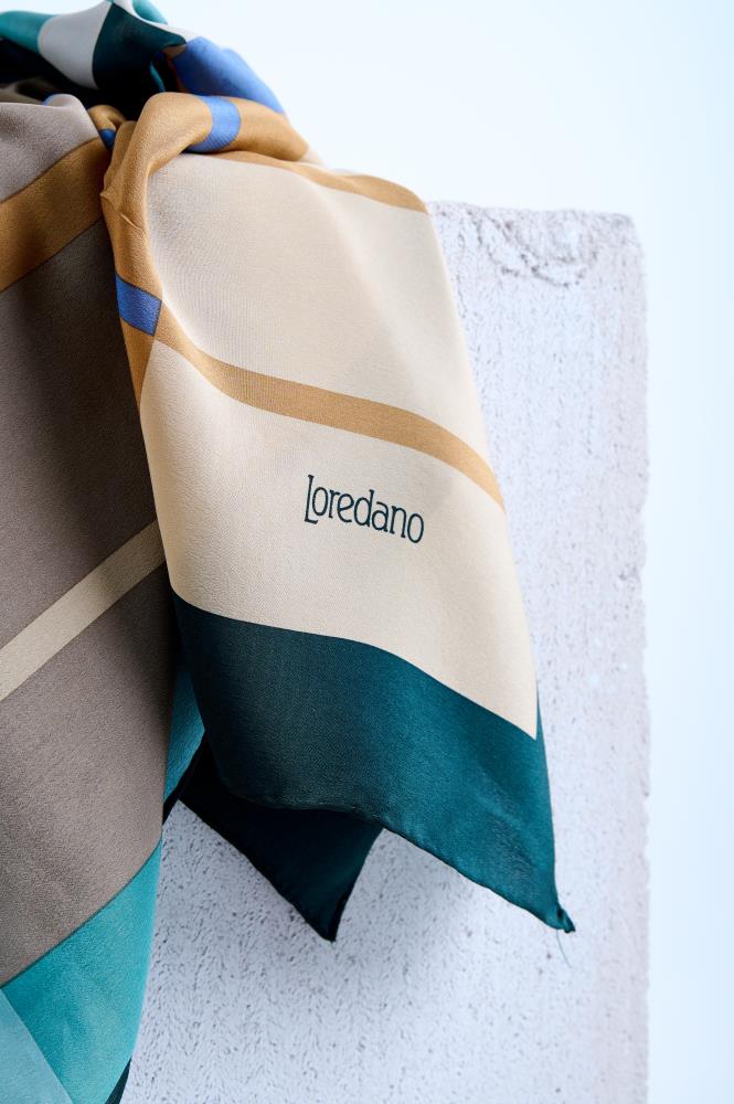 Close-up of a luxury Loredano silk scarf, showcasing the elegant brand logo and a modern geometric stripe pattern in cream, tan, and deep teal, draped over a textured white surface.