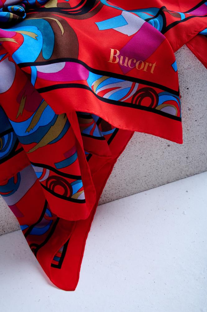 Close-up of a vibrant red silk scarf: a modern abstract print with teal, magenta, and brown geometric patterns, draped over a textured stone edge.
